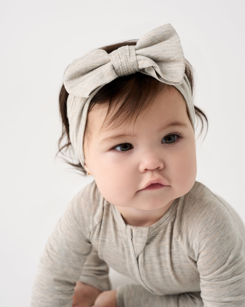 A baby wearing a light beige heathered long-sleeve bodysuit and a matching fabric bow headband, photographed against a clean white studio background.