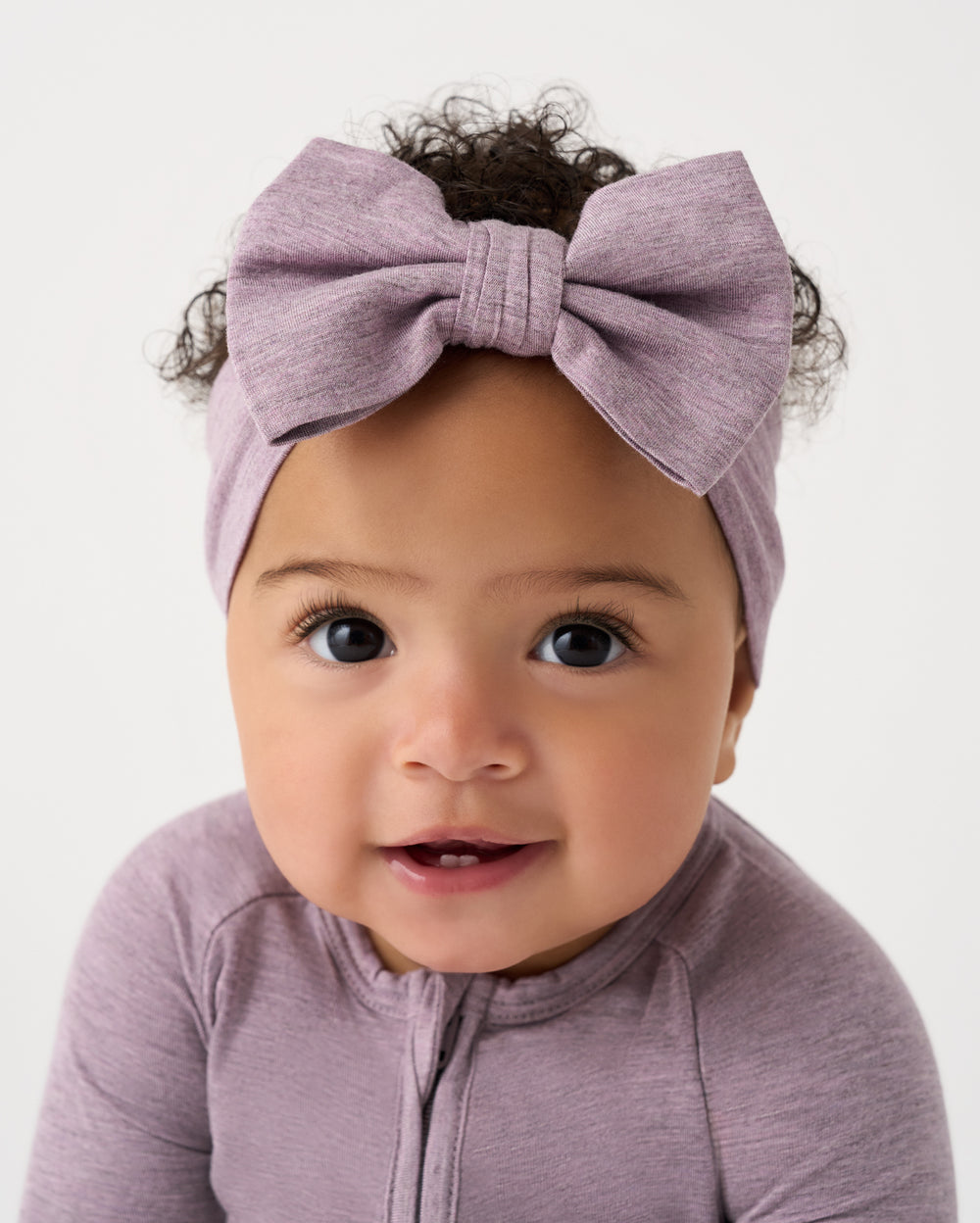 A close-up of a baby wearing a large fabric bow headband and a matching long-sleeve top in a dusty mauve-purple color, set against a plain light gray background.