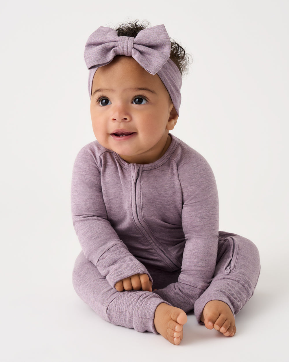 A baby wearing a heathered dusty purple long-sleeve zip-up one-piece and a matching bow headband, sitting against a plain white studio background.