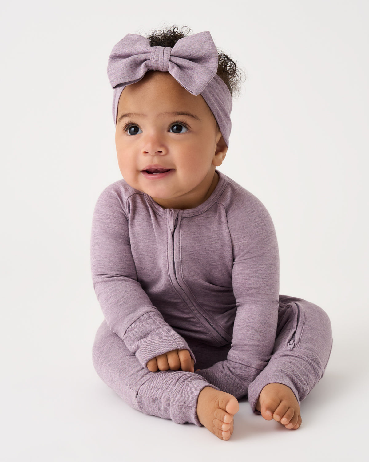 A baby wearing a heathered dusty purple long-sleeve zip-up one-piece and a matching bow headband, sitting against a plain white studio background.