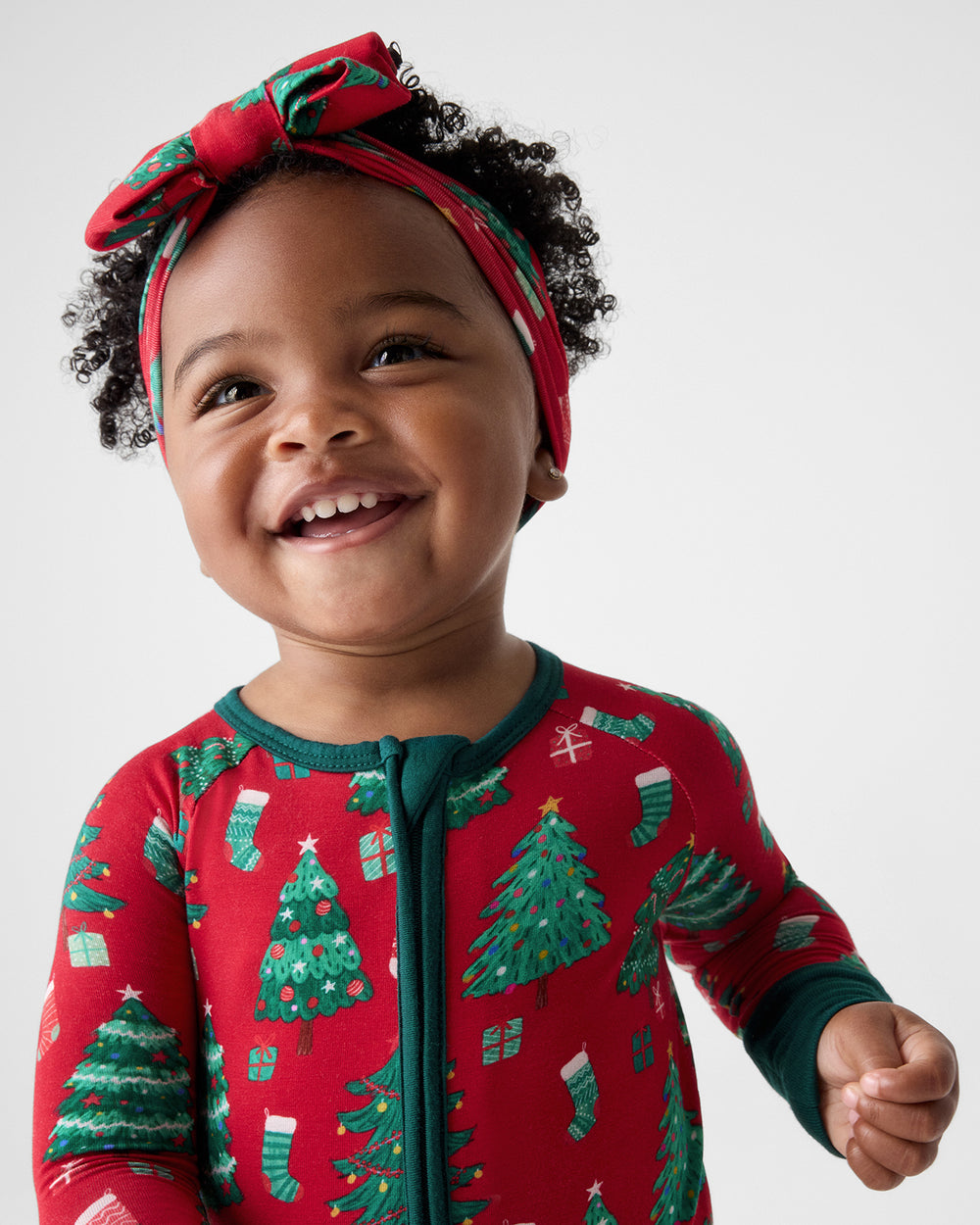 A smiling toddler wearing a festive red and green Christmas print zip-up pajama and a matching red bow headband, photographed against a clean light gray studio background.