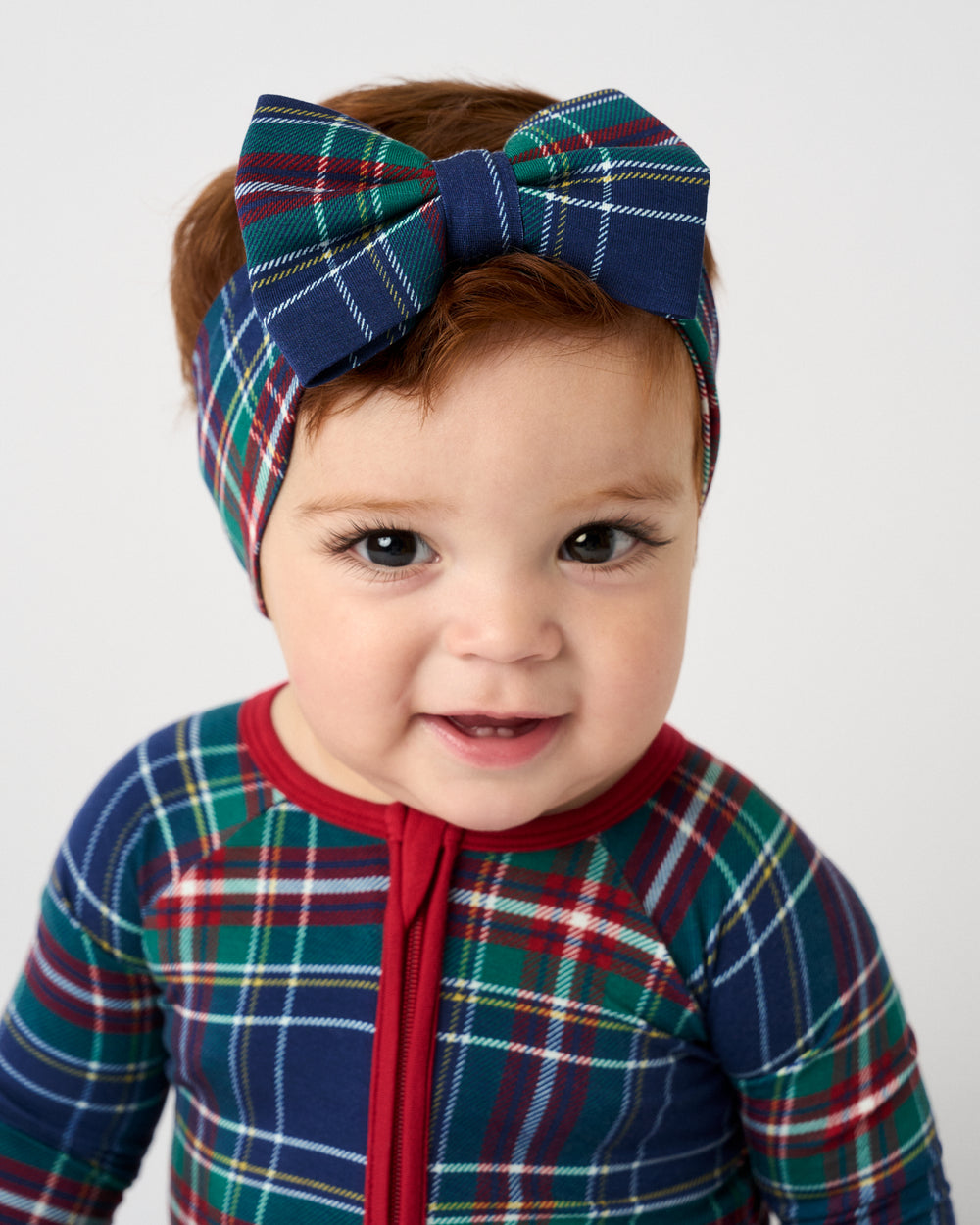A baby wearing a matching plaid one-piece outfit and a large plaid bow headband, photographed against a clean light gray background.