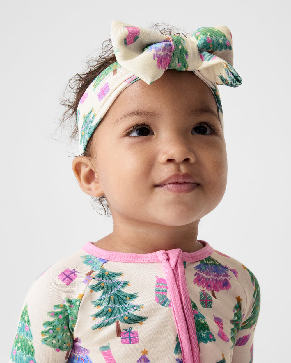 A close-up of a young child wearing a cream-colored knit top and matching bow headband, both decorated with a colorful Christmas tree and gift print, against a plain light gray background.