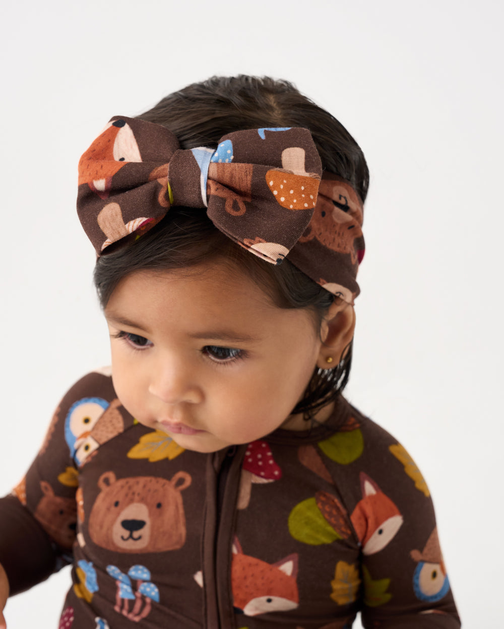 A close-up of a toddler wearing a brown zip-front one-piece outfit and matching bow headband, both printed with a woodland animal pattern including bears, foxes, owls, and leaves on a light gray background.