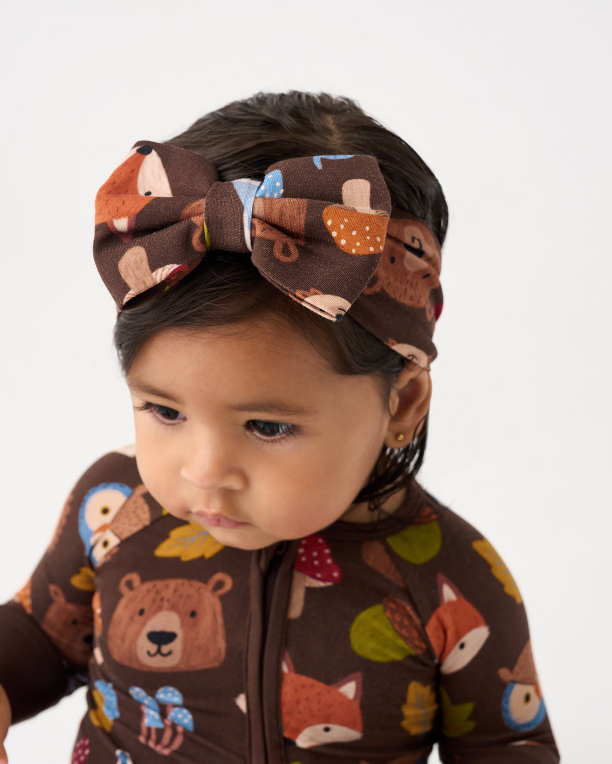 A close-up of a toddler wearing a brown zip-front one-piece outfit and matching bow headband, both printed with a woodland animal pattern including bears, foxes, owls, and leaves on a light gray background.