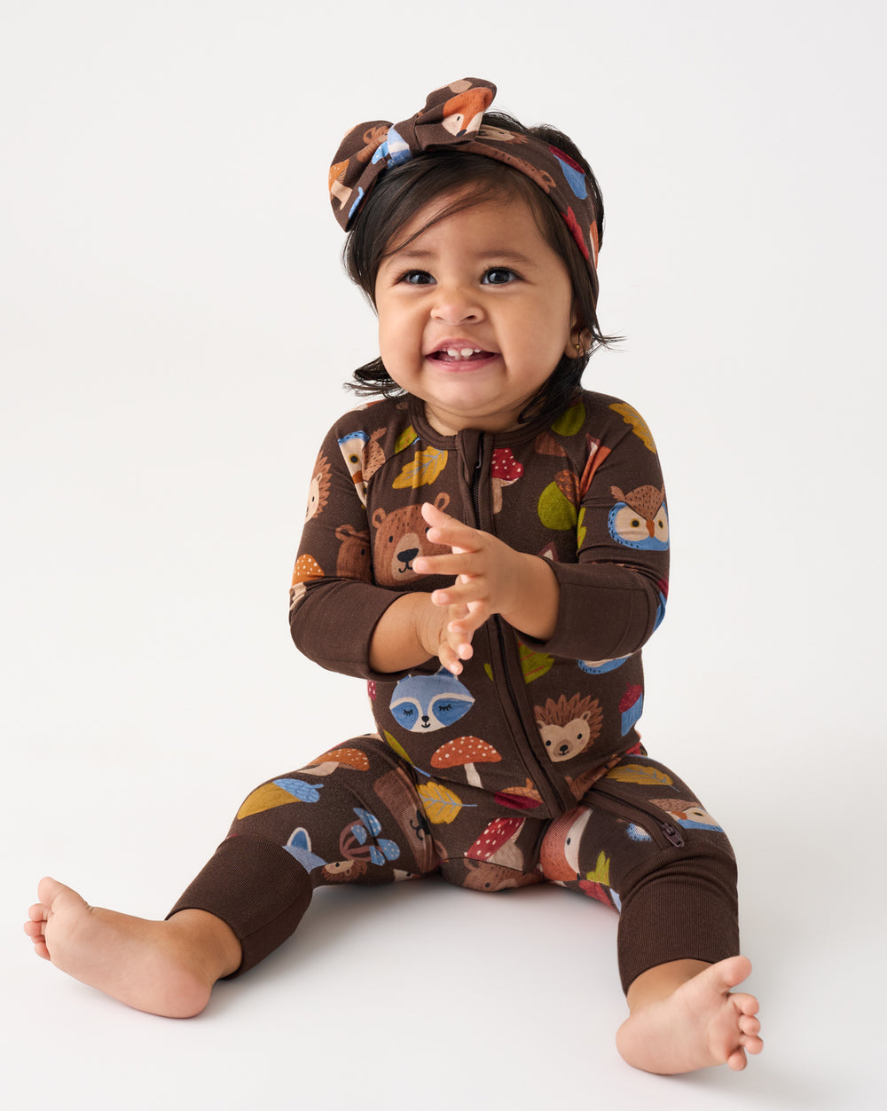 A baby sitting on a white studio floor wearing a brown zip-front one-piece sleeper with colorful woodland animal prints and a matching headband.