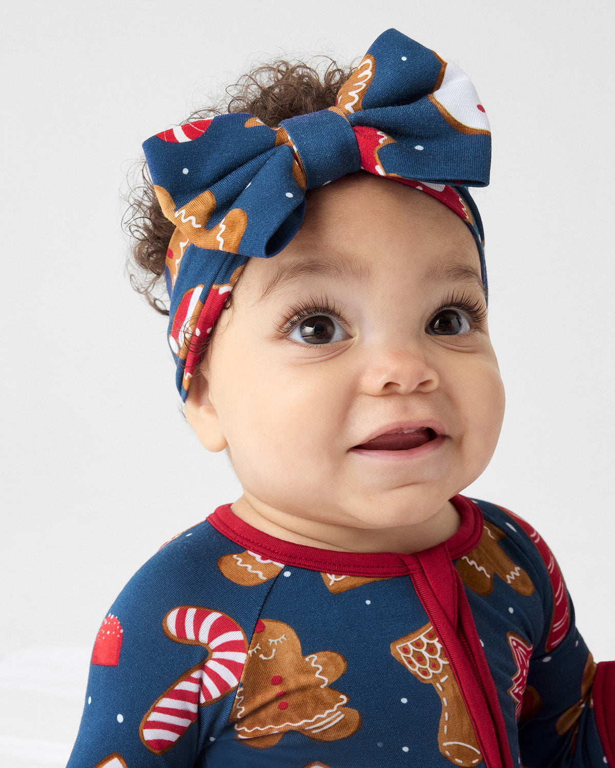 A baby wearing a navy blue holiday-themed PJ with gingerbread and candy cane prints, paired with a matching fabric bow headband, photographed against a light gray background.