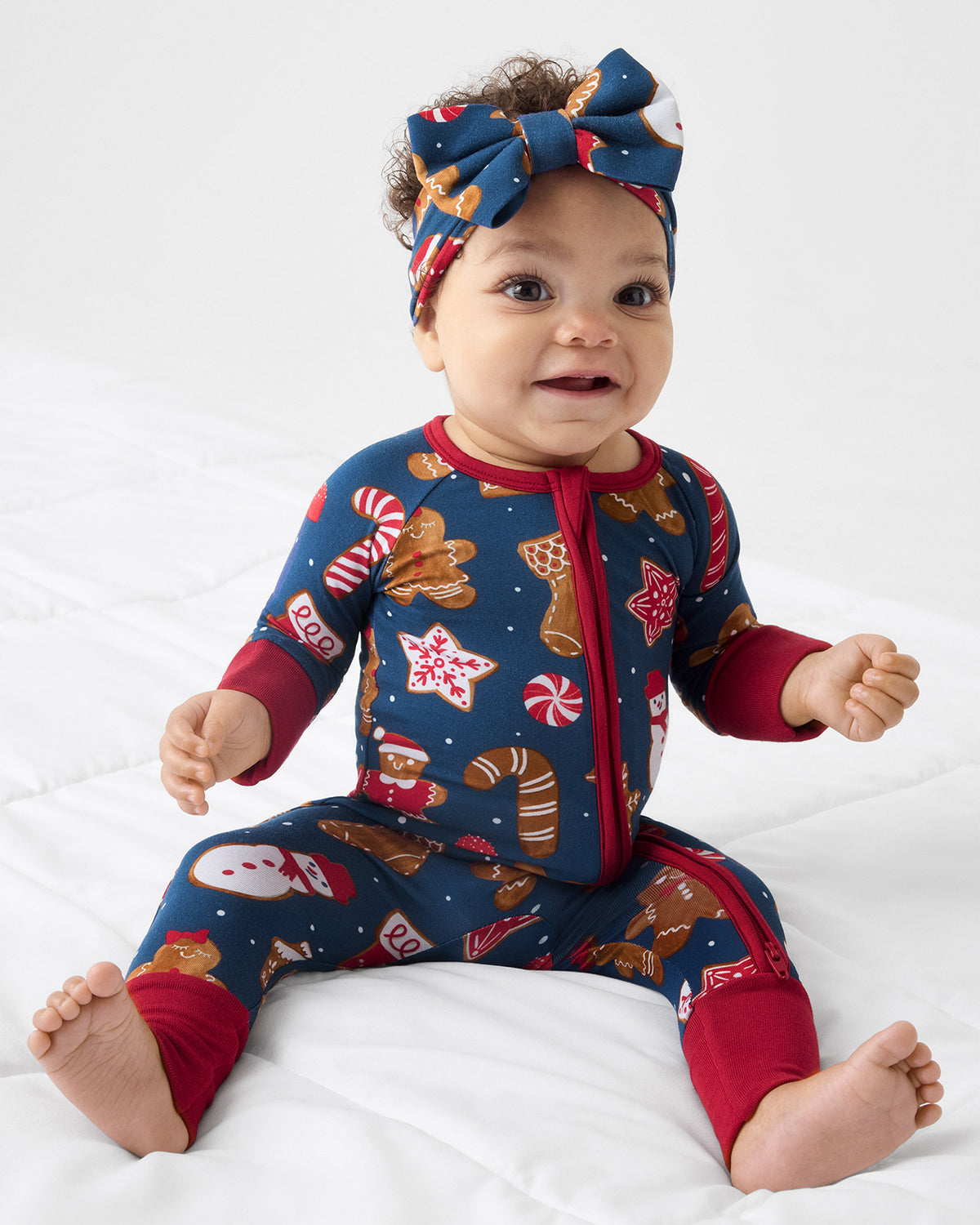 A baby sitting on a white quilted bed wearing a festive Christmas-themed one-piece PJ with a matching bow headband. The Zippy features a navy blue background decorated with gingerbread cookies, candy canes, snowman prints, and red cuffs and zipper trim.