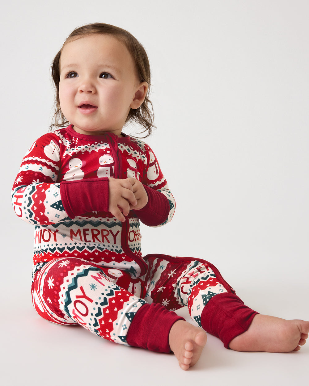 A baby sitting on a light gray studio background wearing a red, white, and navy Christmas-themed one-piece pajama with 'MERRY JOY' text and holiday motifs.