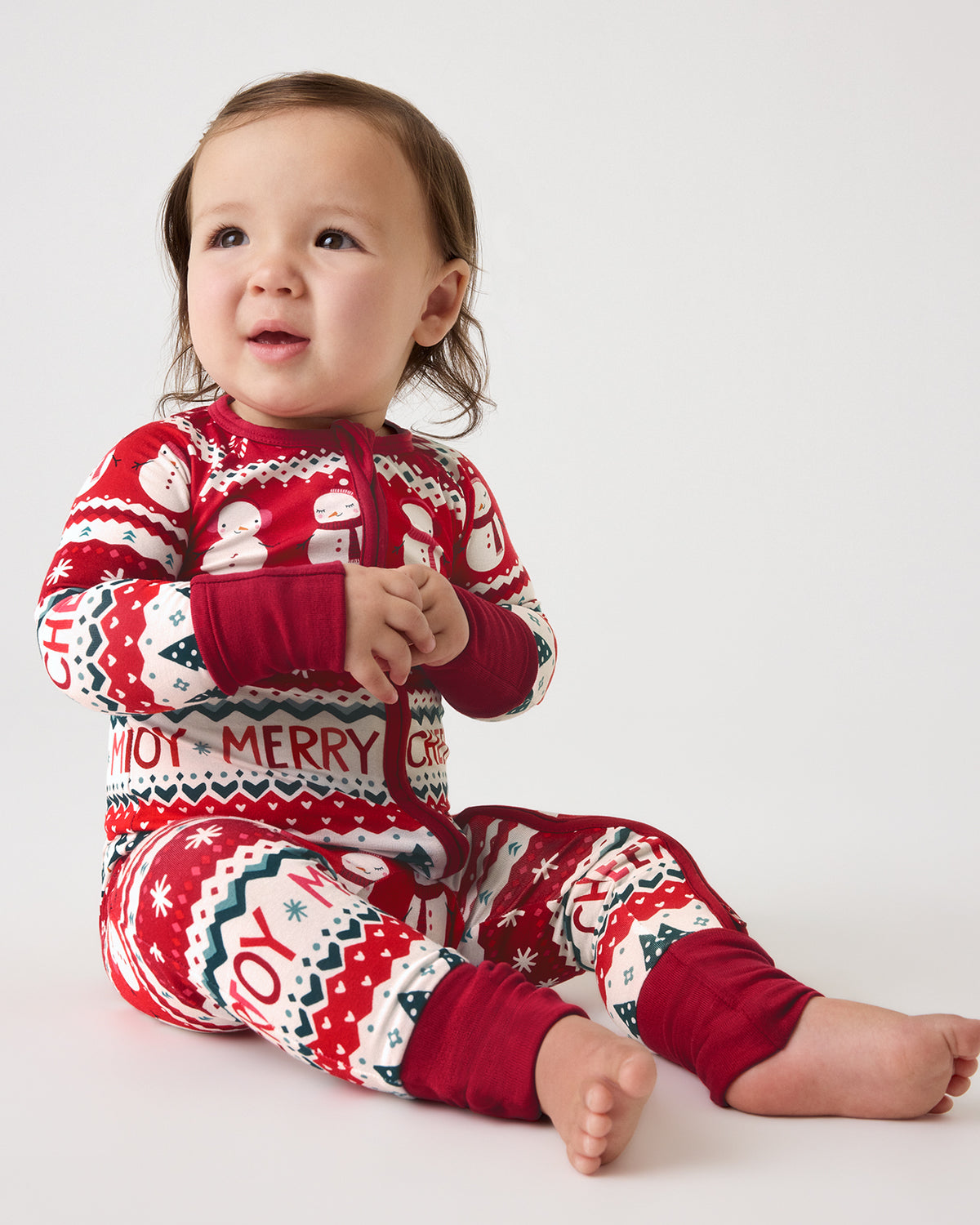 A baby sitting on a light gray studio background wearing a red, white, and navy Christmas-themed one-piece pajama with 'MERRY JOY' text and holiday motifs.