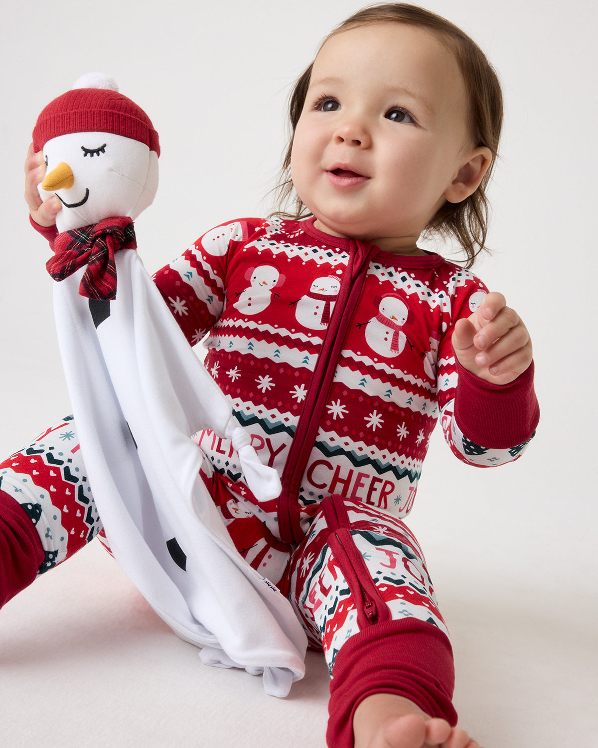 A baby wearing a red and white Christmas-themed zip-up one-piece pajama sits on a light studio background while holding a plush snowman toy.