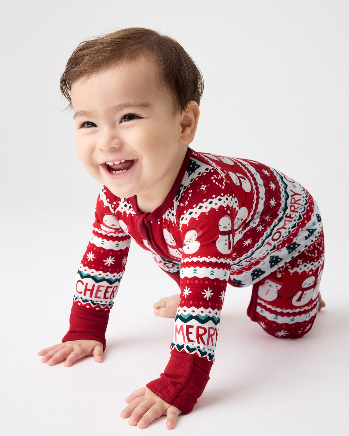 A smiling baby crawling on a white studio floor wearing a red, white, and green Christmas-themed long-sleeve footless pajama top with snowman and 'MERRY' patterns.