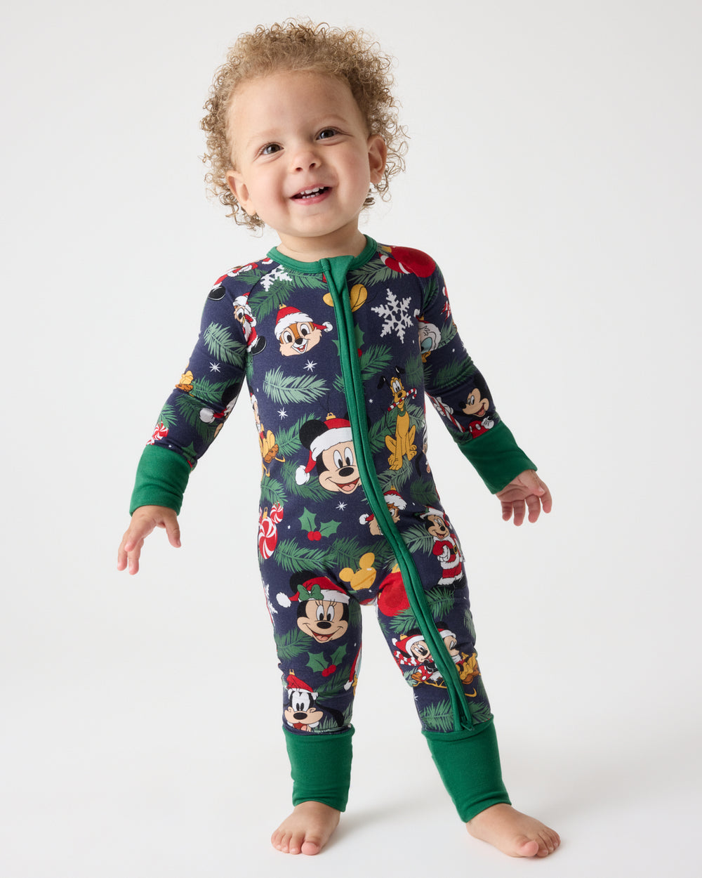 A toddler standing barefoot against a light gray studio background, wearing a long-sleeve zip-front pajama one-piece covered in a Christmas-themed Mickey Mouse print with dark green cuffs and collar.