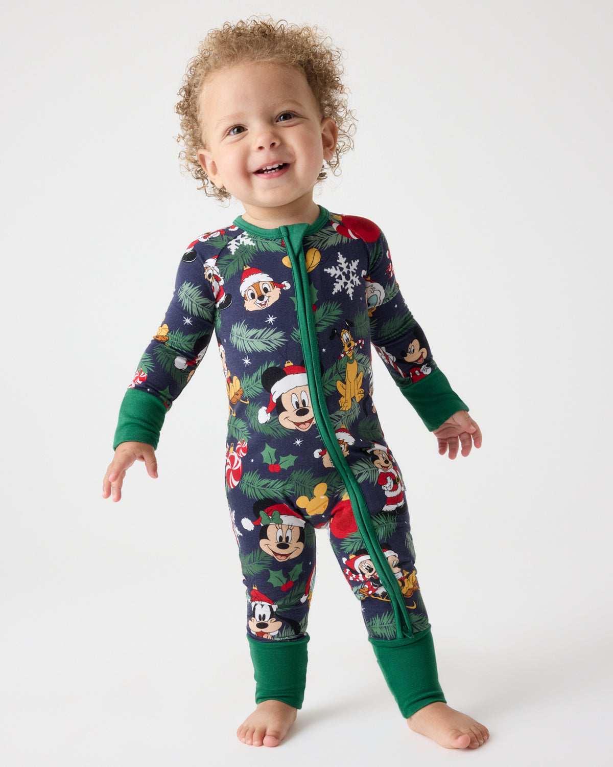 A toddler standing barefoot against a light gray studio background, wearing a long-sleeve zip-front pajama one-piece covered in a Christmas-themed Mickey Mouse print with dark green cuffs and collar.
