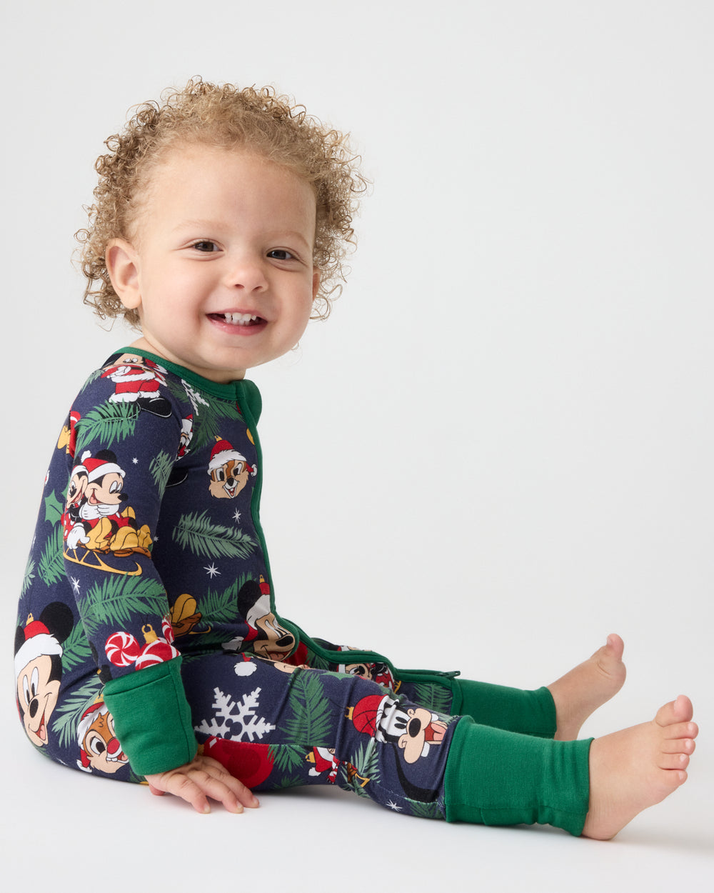 A smiling toddler sits on a light gray studio floor wearing a festive Christmas-themed one-piece pajama set with Mickey Mouse and Minnie Mouse prints, green cuffs, and a dark navy base.