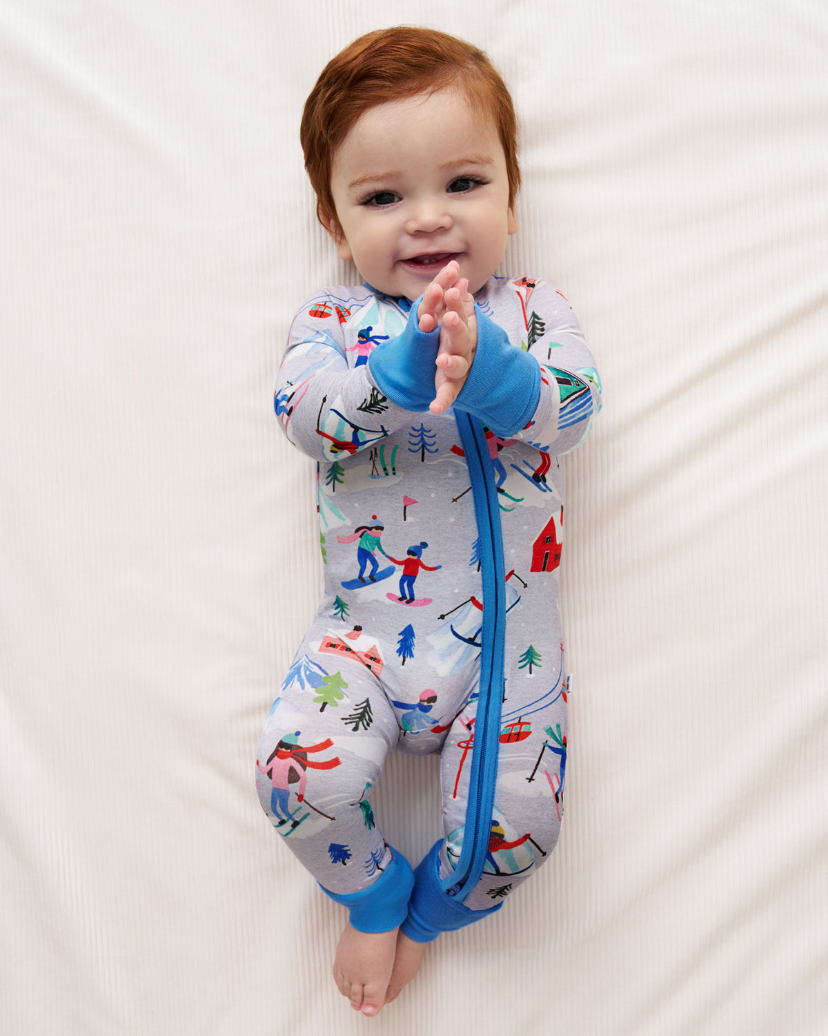 A baby with red hair lies on a white textured sheet, wearing a light gray footed sleeper with a blue zipper and cuffs, and a colorful winter ski pattern.