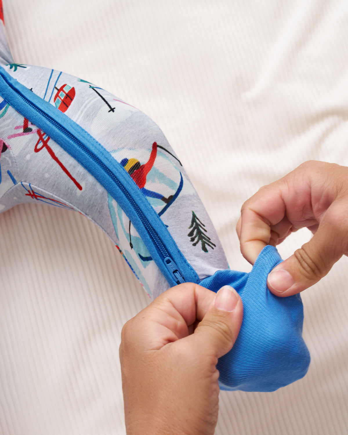 Close-up of a person's hands attaching a bright blue fabric pouch to a curved, ski-patterned support pillow on a white ribbed fabric surface.