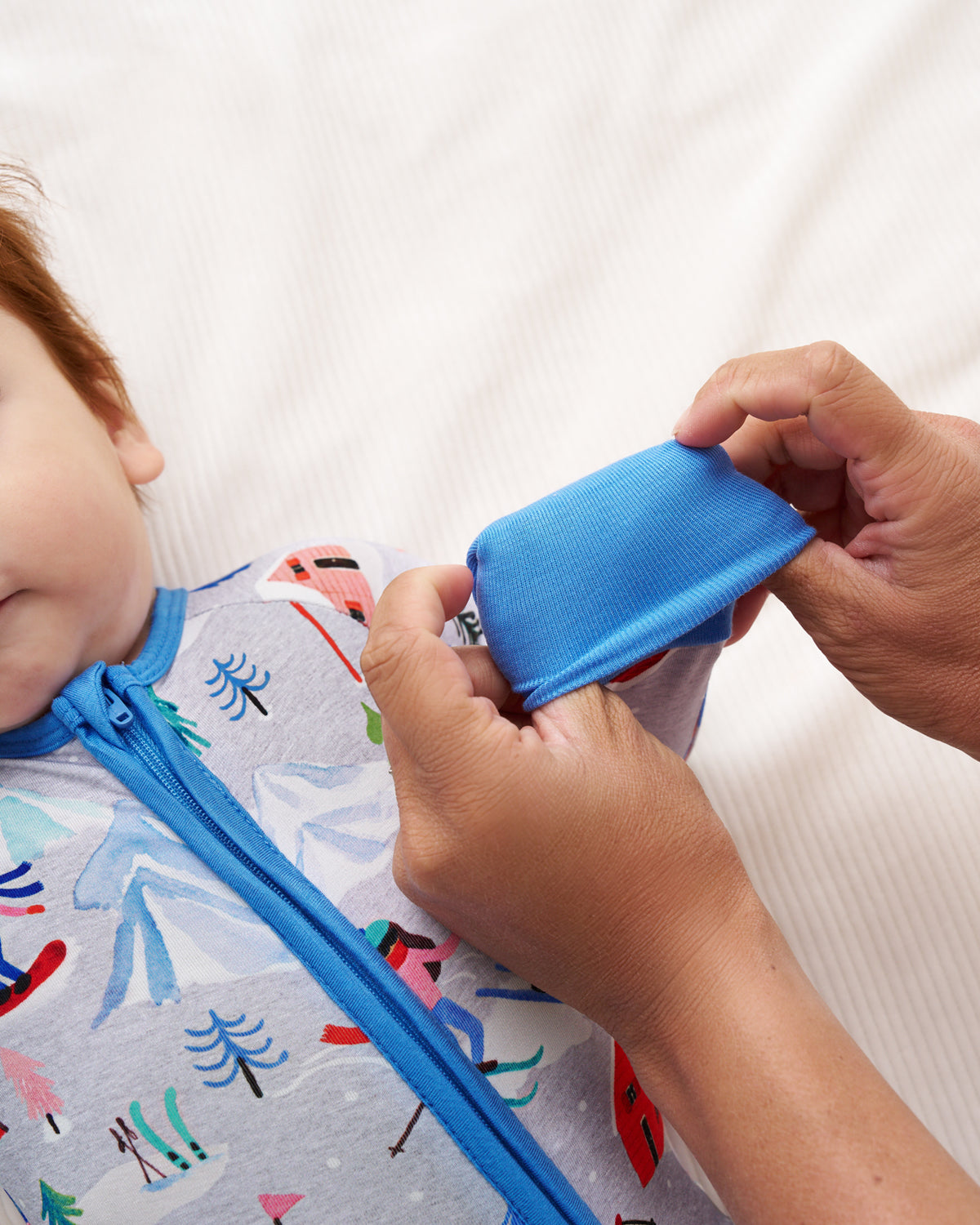 A close-up of a baby wearing a light gray patterned sleep sack with a blue zipper, while an adult's hands adjust a matching blue neck warmer or hood on the baby.