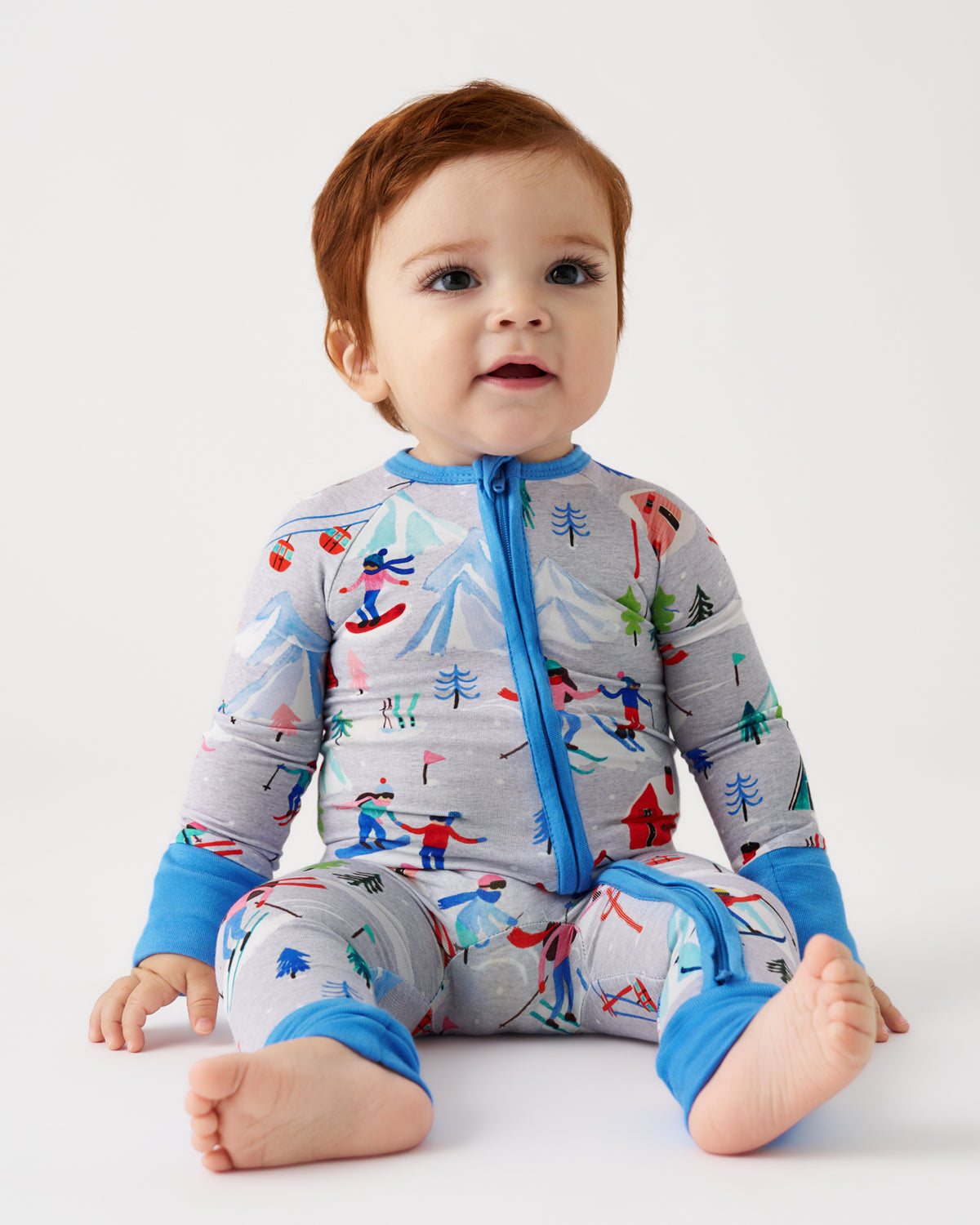 A baby sitting on a white studio floor wearing a long-sleeve zip-up sleeper with a gray background and a colorful alpine ski pattern.