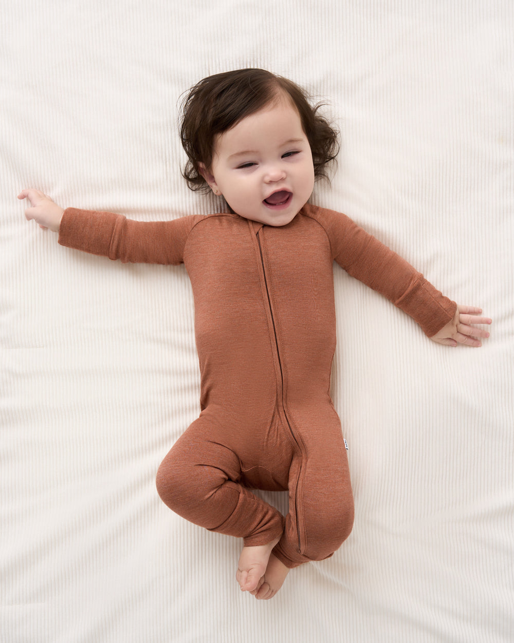 A baby lying on a white textured blanket wearing a rust-brown long-sleeve zip-front footed sleeper.