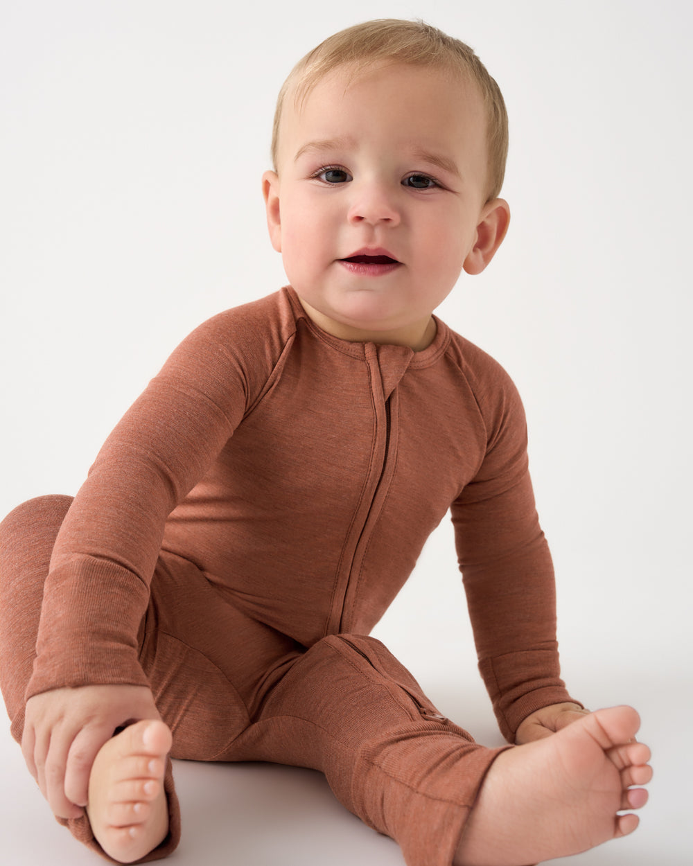 A baby wearing a rust-brown long-sleeve zip-up footed one-piece, sitting against a plain white studio background.