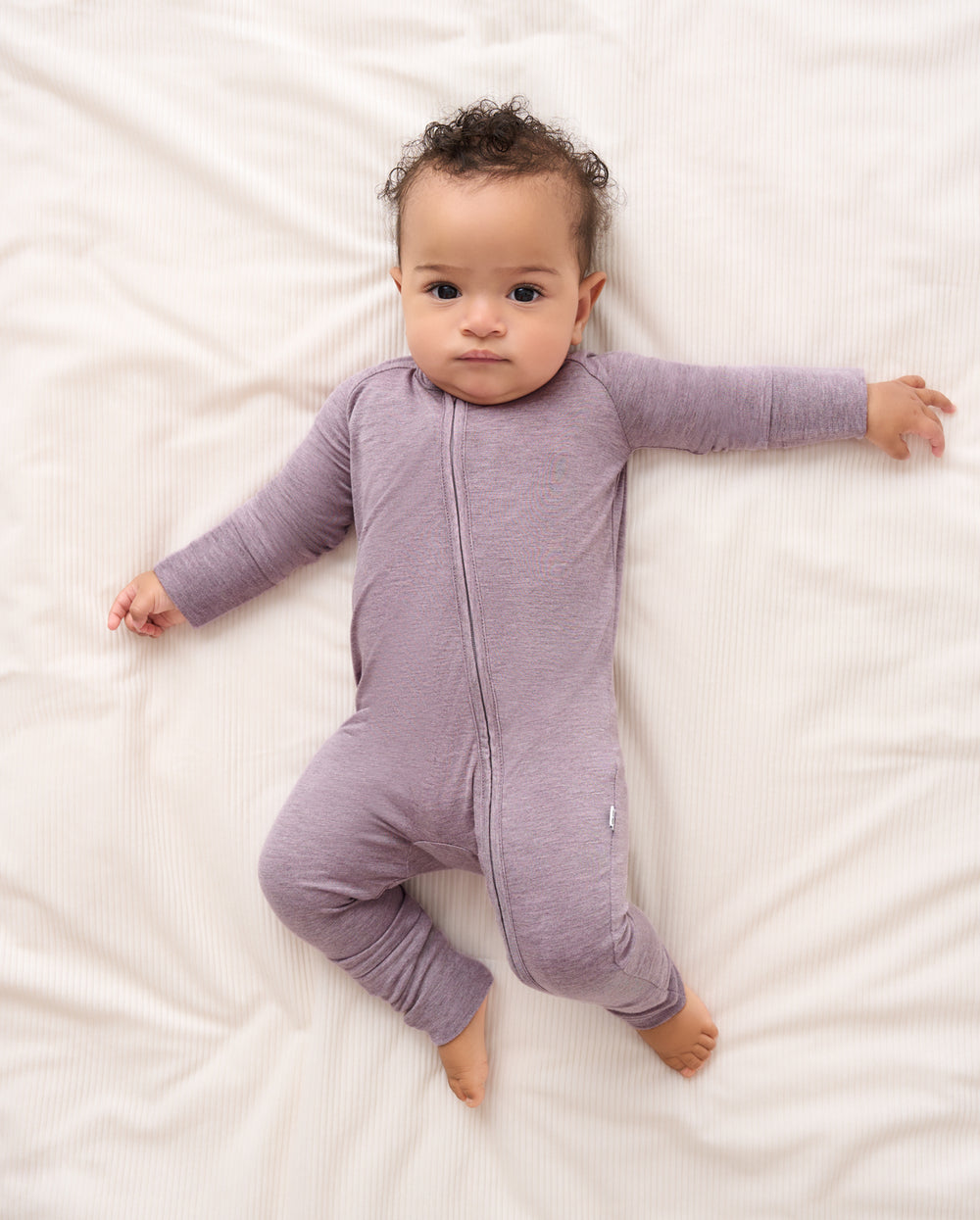 A baby lying on white bedding wearing a heathered light purple long-sleeve footless sleeper with a front zipper.
