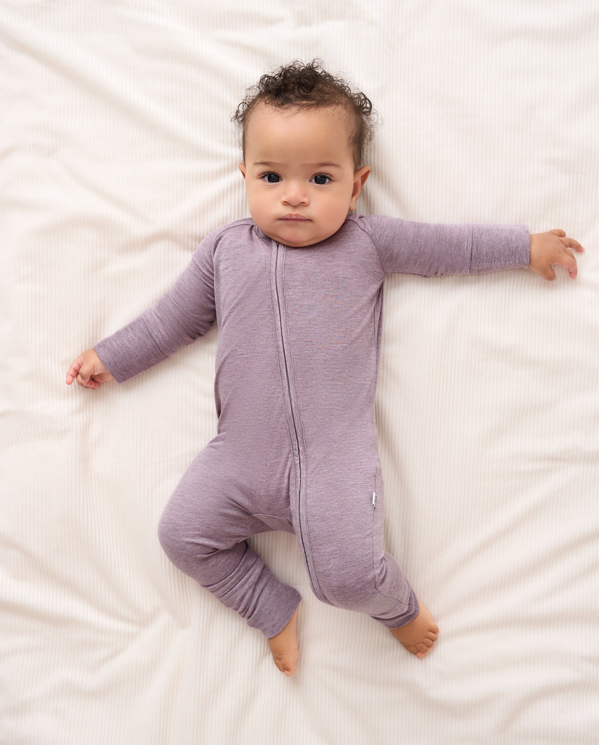 A baby lying on white bedding wearing a heathered light purple long-sleeve footless sleeper with a front zipper.