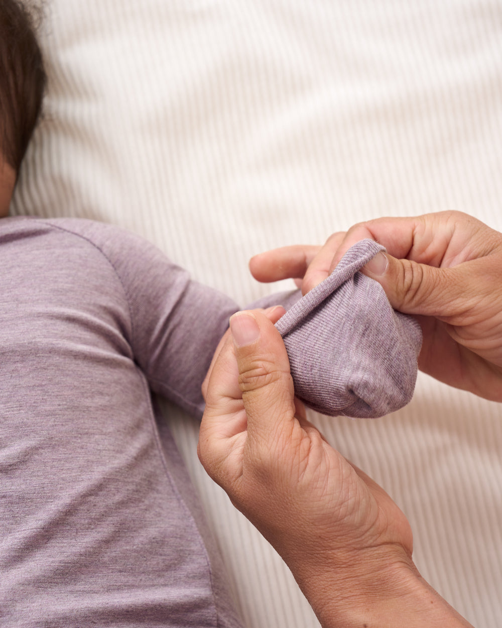 A close-up of a person's hands folding a light purple knit sleeve, with another person wearing a matching light purple long-sleeve top lying down in the background.