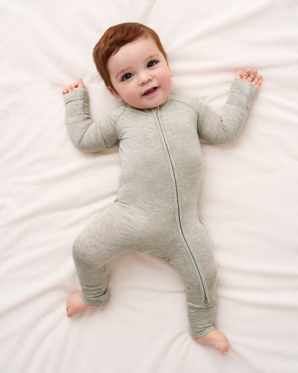 A baby lying on a white textured blanket wearing a light gray heathered zip-up sleeper.