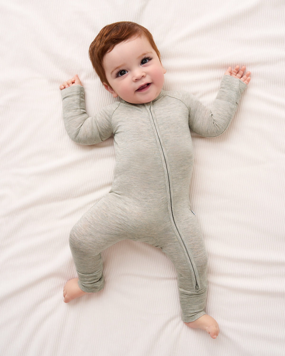 A baby lying on a white textured blanket wearing a light gray heathered zip-up sleeper.