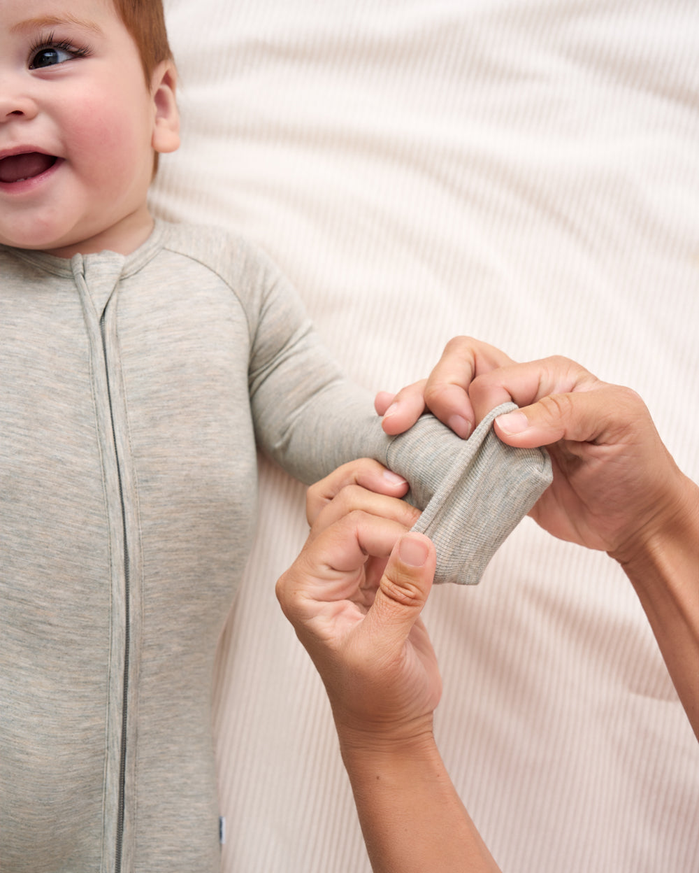 A baby wearing a light gray long-sleeve zip-up sleeper, with an adult's hands adjusting the sleeve. The baby is lying on a white textured blanket.