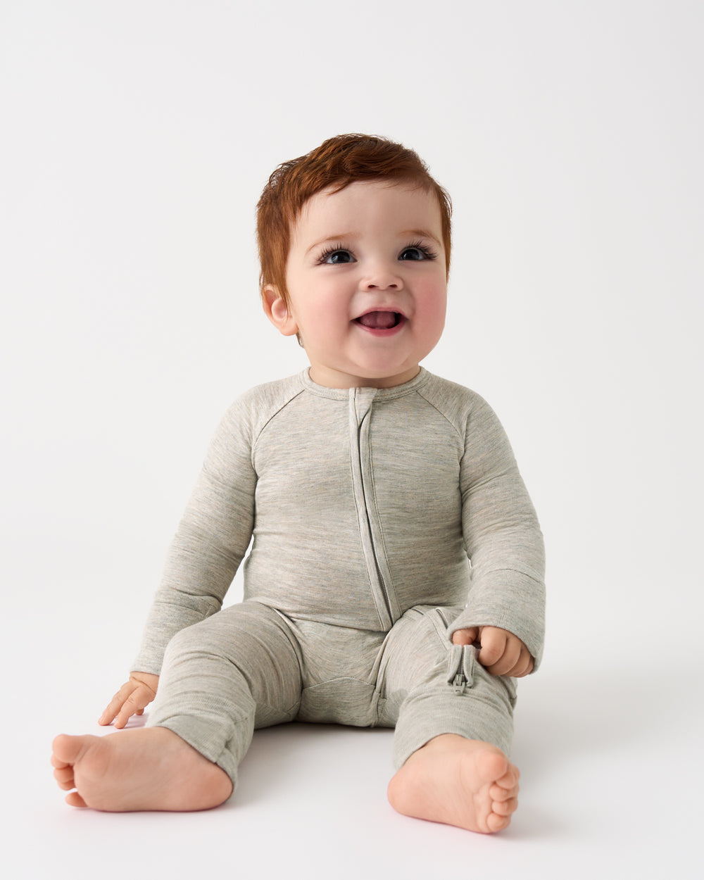 A smiling baby with short brown hair sits on a white studio floor wearing a light heather gray long-sleeve zip-front footless sleeper.