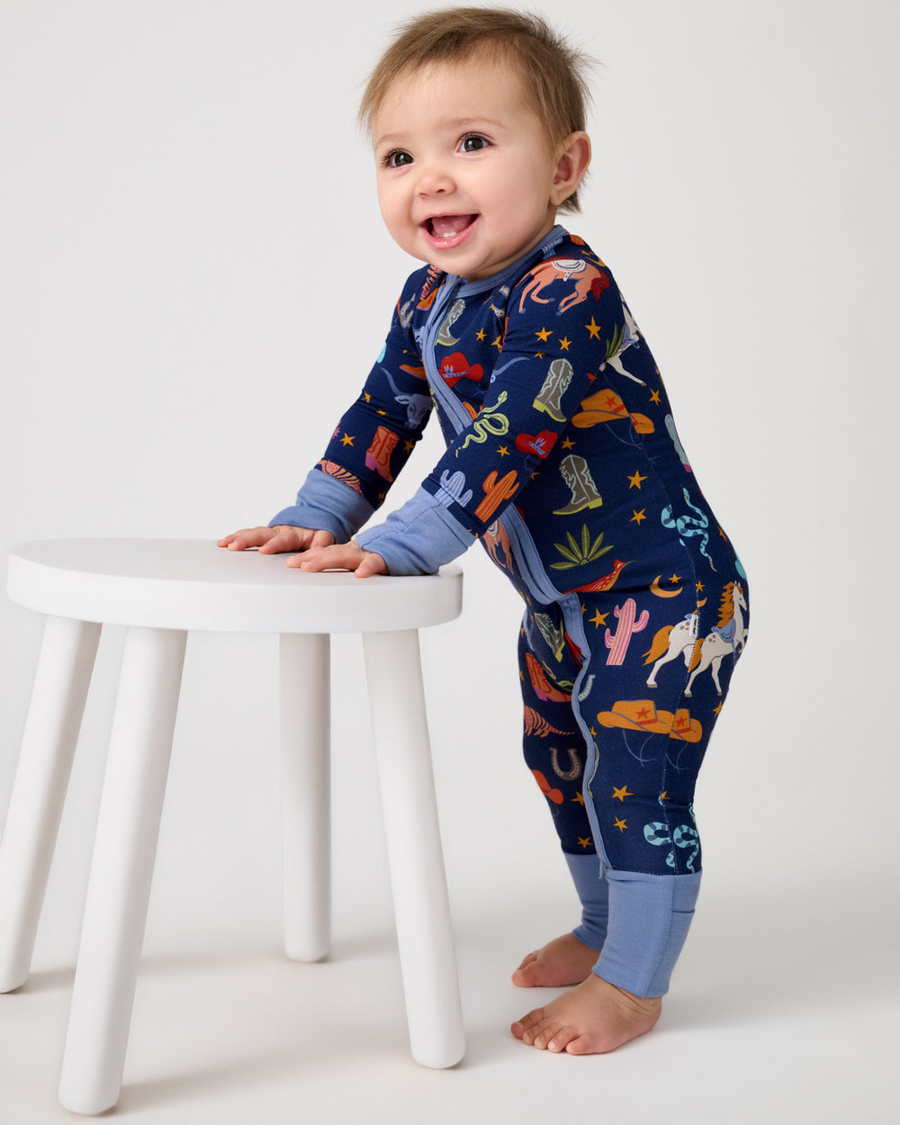 Baby holding onto a white stool wearing the Blue Rancher Roundup Zippy
