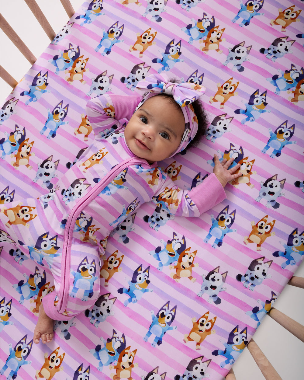 Baby laying on the Pink Bluey Dance All Day Fitted Crib Sheet in a matching zippy