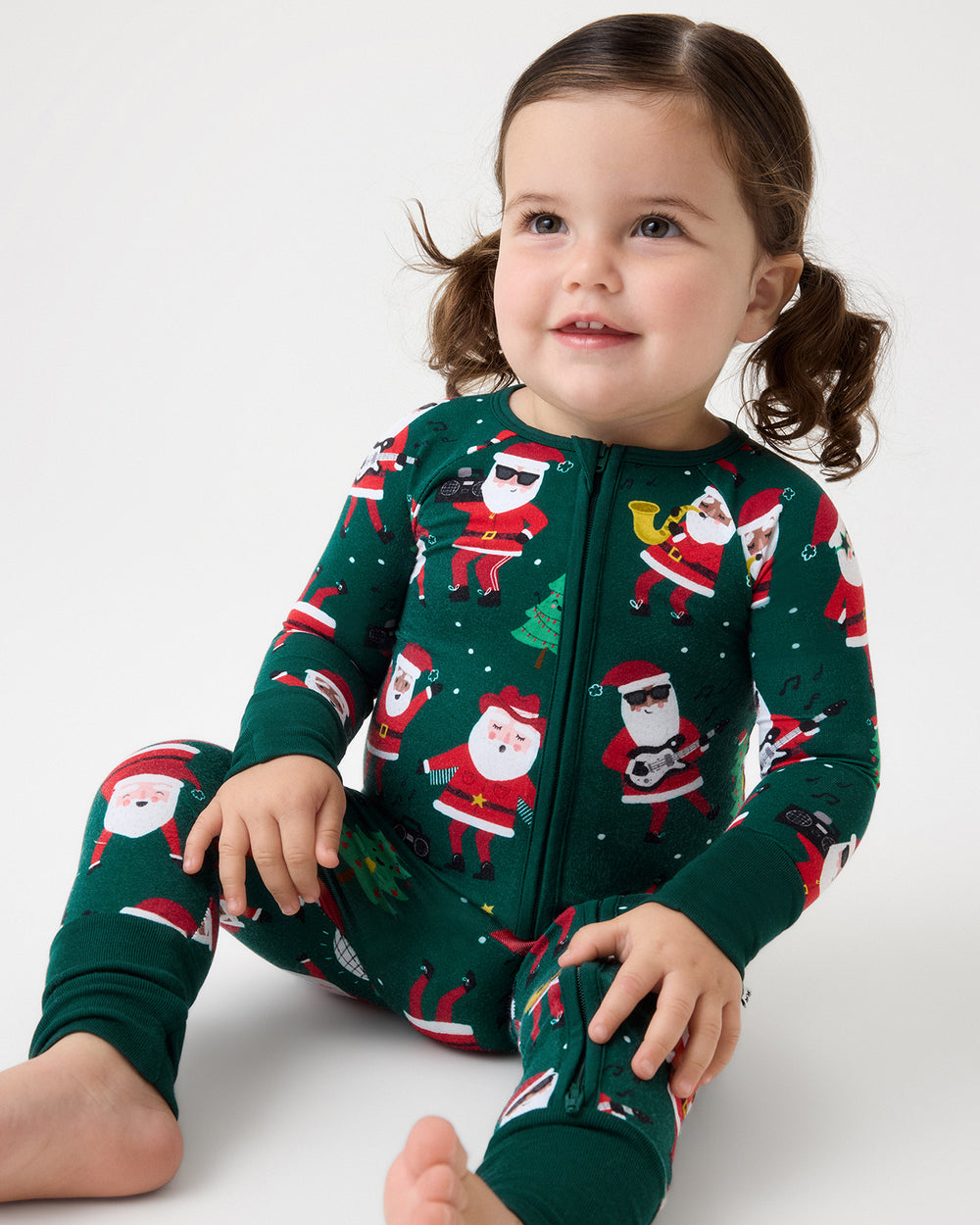 A young child wearing a dark green full-body zip-up pajama with a festive Santa Claus print, sitting against a plain light gray studio background.