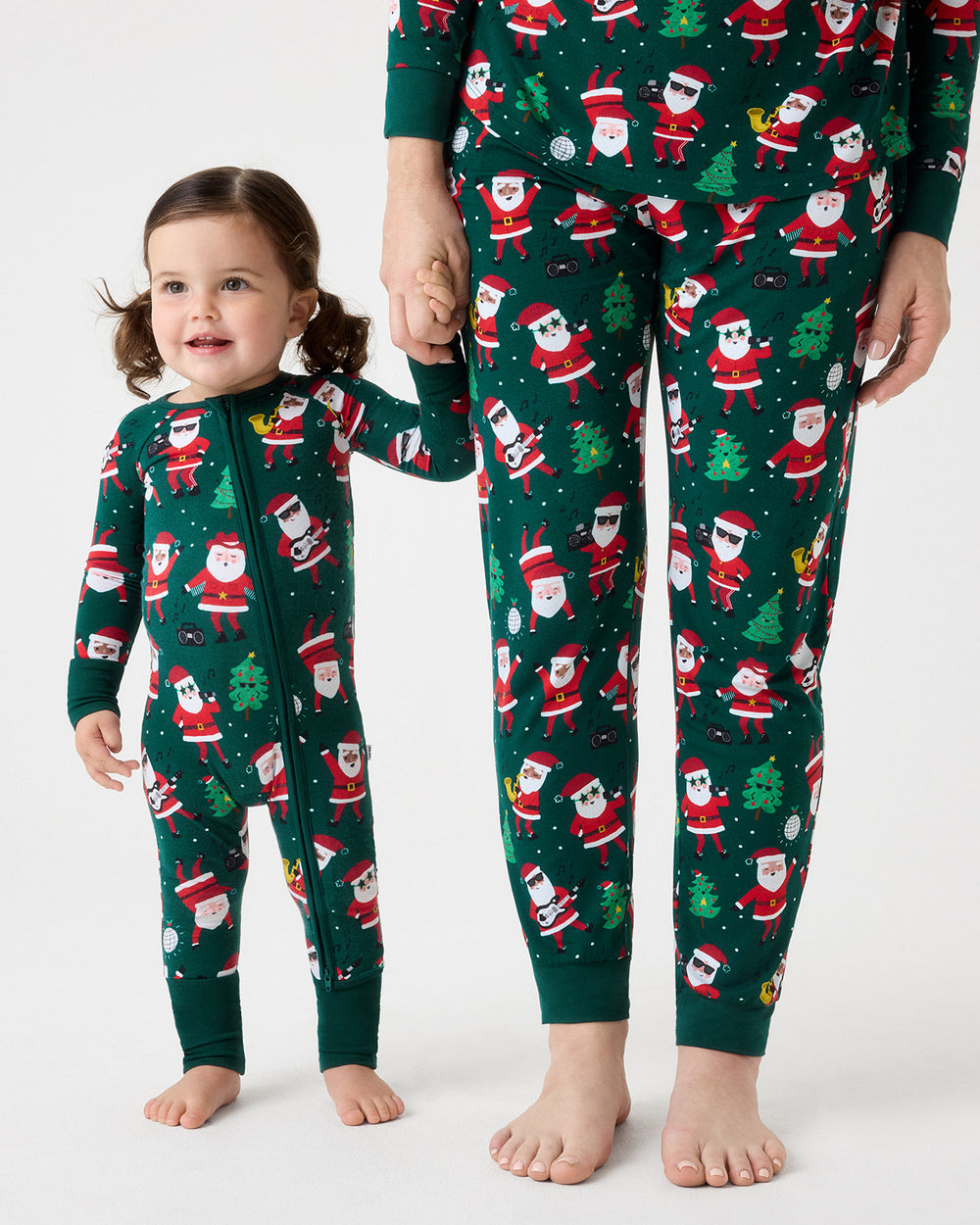 A child wearing a green Christmas-themed zip-up one-piece pajama holds hands with an adult wearing matching green pajama pants and top, both featuring a Santa Claus and Christmas tree pattern on a white studio background.