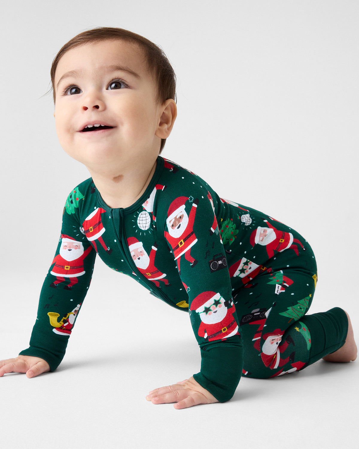 A baby crawling on a light gray studio floor wearing a dark green long-sleeve footie pajama with a festive Santa Claus print.
