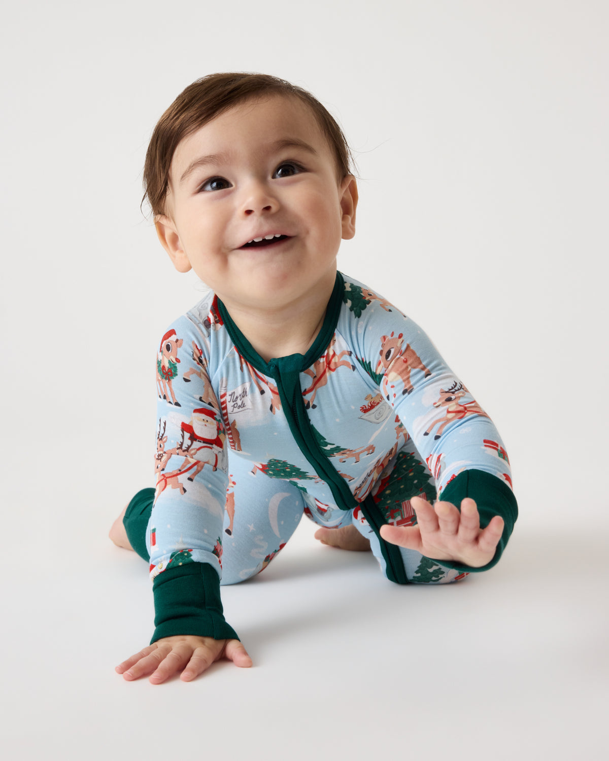 A smiling baby crawling on a white studio floor, wearing a light blue long-sleeve pajama with a Christmas tree and holiday print, dark green trim, and dark green cuffs.