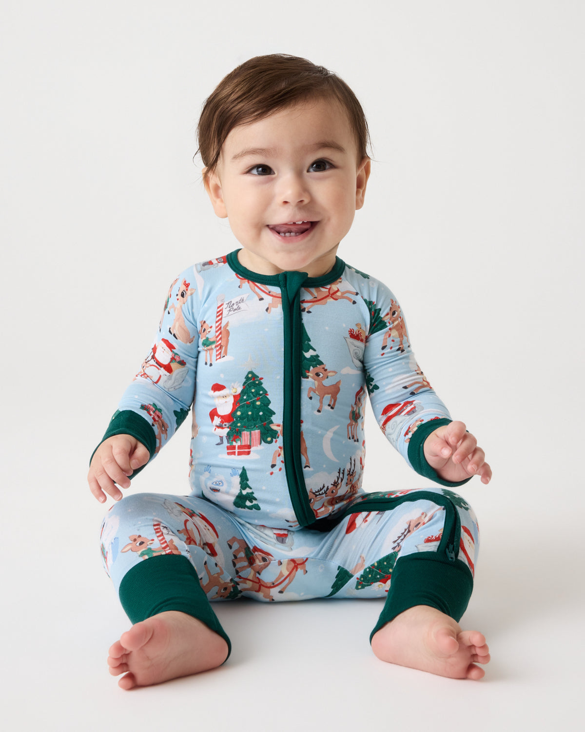 A smiling baby sitting wearing a light blue Christmas-themed long-sleeve sleeper with dark green trim and a festive print of trees, Santa Claus, and reindeer, against a plain white studio background.