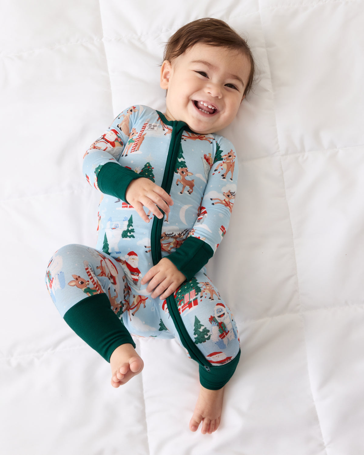 A smiling baby lying on a white quilted blanket, wearing a light blue Christmas-themed footed sleeper with dark green cuffs and zipper trim.