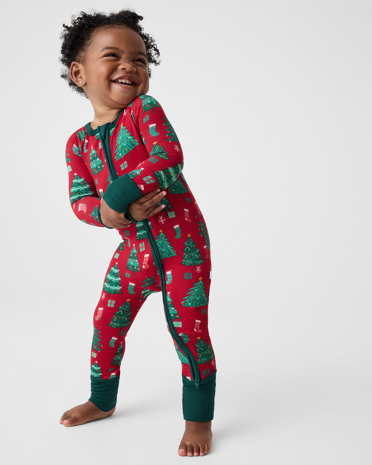 A smiling toddler stands barefoot against a light gray studio background, wearing a one-piece zip-up pajama in red with a green Christmas tree and gift print and dark green cuffs and collar.