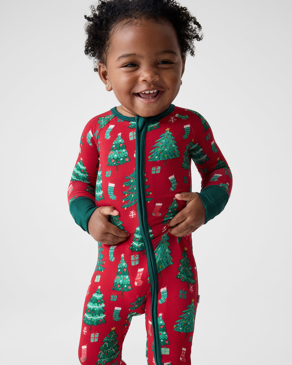 A smiling toddler wearing a red one-piece pajama with a green zipper and a green Christmas tree and stocking print, photographed against a light gray studio background.