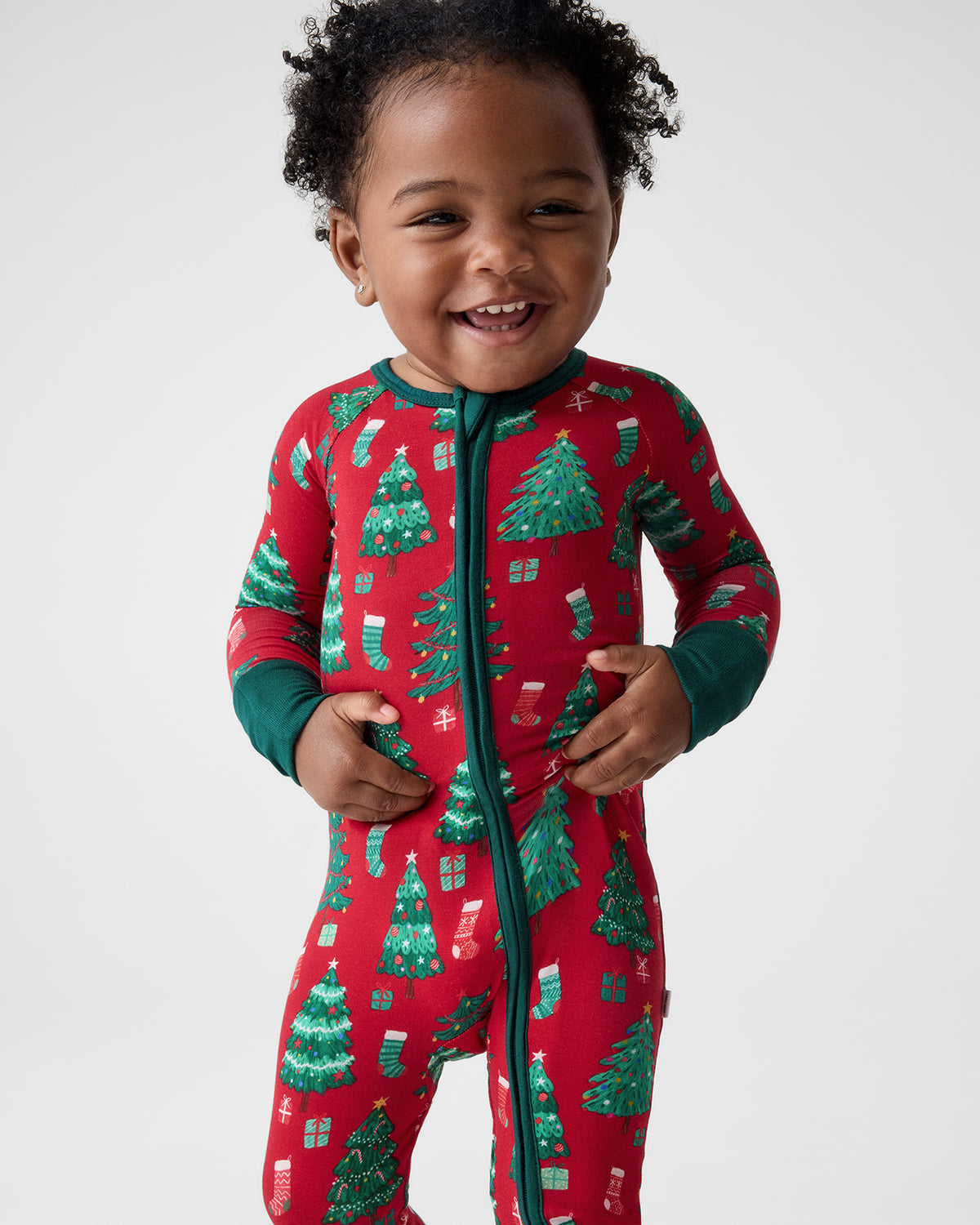 A smiling toddler wearing a red one-piece pajama with a green zipper and a green Christmas tree and stocking print, photographed against a light gray studio background.