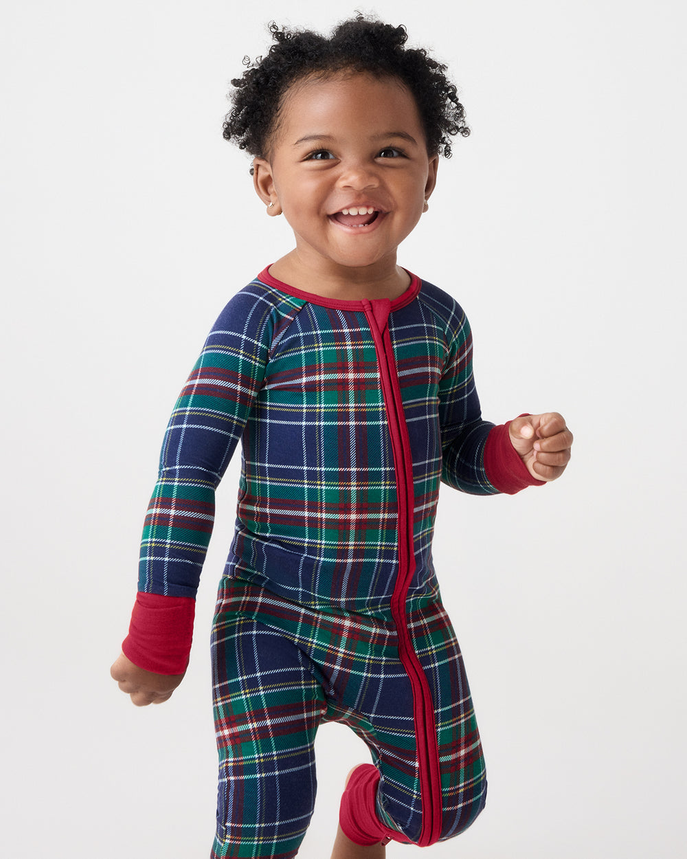 A smiling toddler wearing a long-sleeve footed pajama romper in a navy blue plaid pattern with red, green, and white stripes, featuring a red front zipper and red cuffs, set against a plain light gray background.