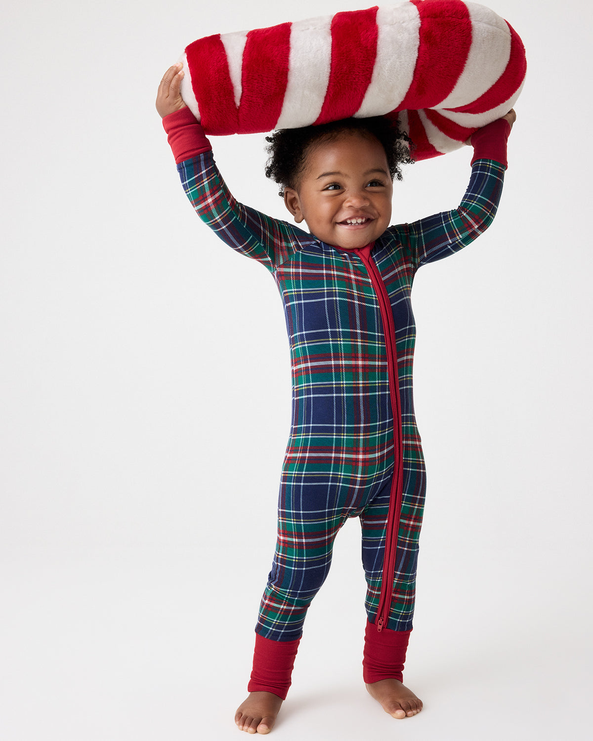 A smiling toddler stands barefoot against a light gray studio background, wearing a full-body zip-up pajama one-piece in a red, green, and navy plaid pattern, with red cuffs and holding a large red-and-white striped pillow overhead.