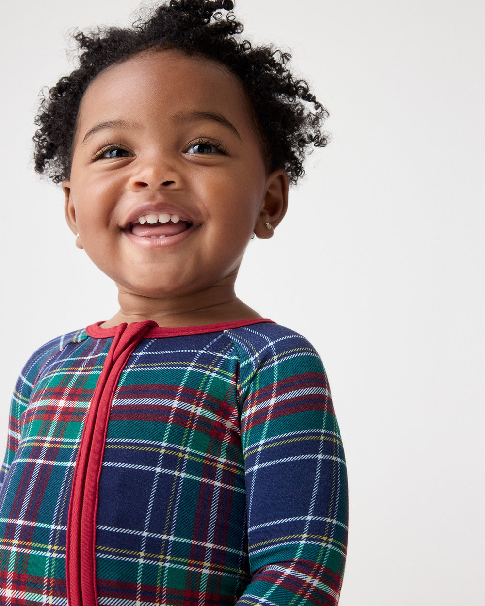 A smiling toddler wearing a long-sleeve plaid zip-up pajama top in navy blue with red, green, and white tartan lines, photographed against a clean light gray background.