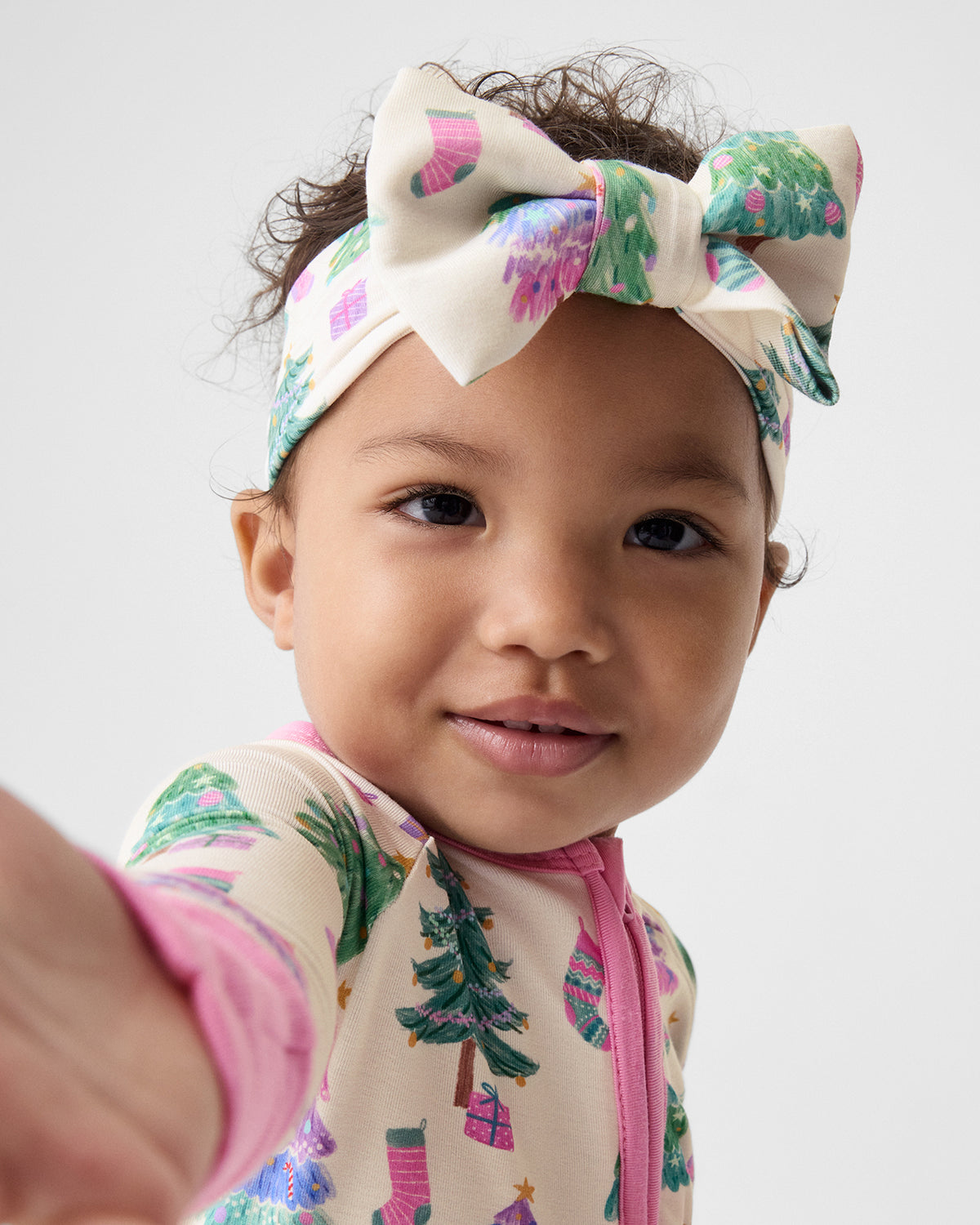 A close-up of a young child wearing a cream-colored pajama top with a pink zipper and a Christmas tree print, paired with a matching cream bow headband featuring a colorful holiday pattern.