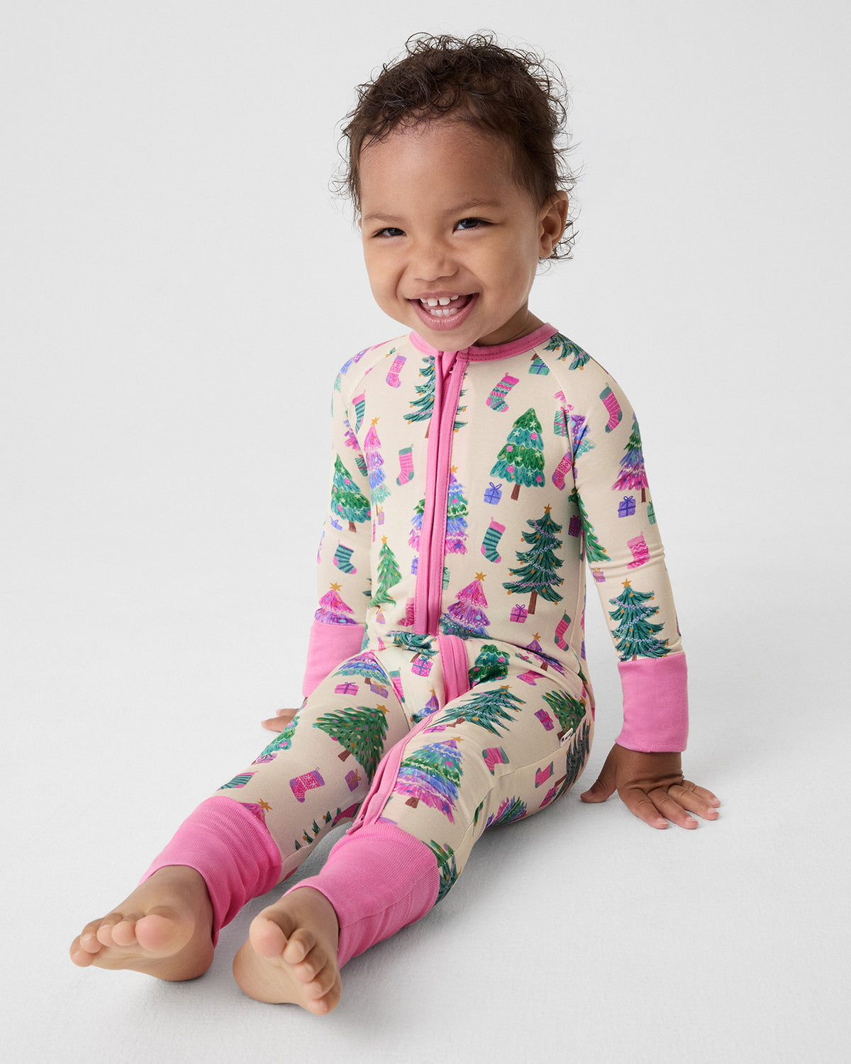 A smiling toddler sits on a light gray studio floor wearing a one-piece zip-up pajama with a cream base and a colorful Christmas tree print, pink cuffs, and a pink zipper.