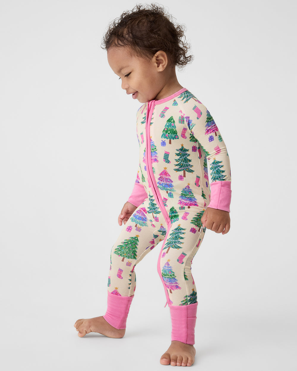 A toddler wearing a long-sleeve zip-front pajama one-piece with a Christmas tree and gift print on a cream background, featuring bright pink cuffs and hem, standing against a light gray studio background.
