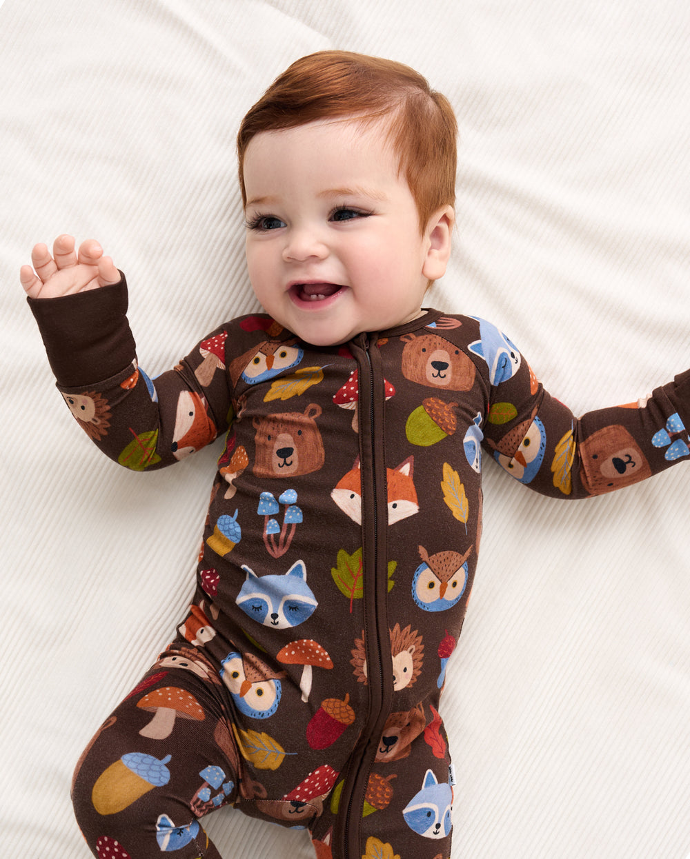 A baby lying on a white textured blanket, wearing a brown long-sleeve footless zip-up sleeper covered in a colorful woodland animal print including foxes, raccoons, owls, mushrooms, and acorns.